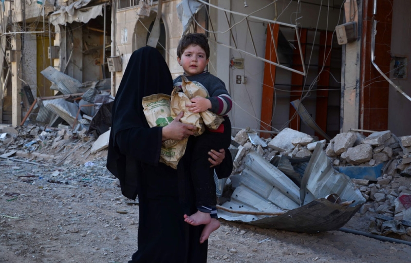 امرأة سورية تحمل طفلا وحزمة من المساعدات الإنسانية في الباب شمال البلاد (أ.ف.ب)