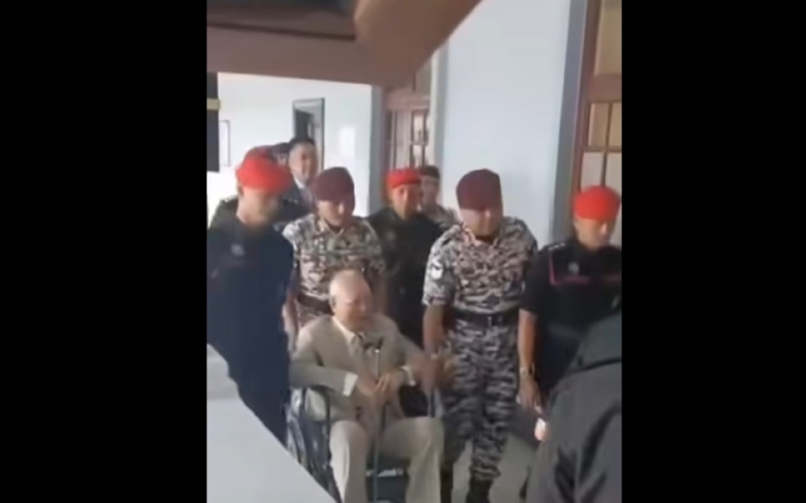 纳吉在狱警的协助下,坐着轮椅到法庭面审。-图截自影片- 纳吉在狱警的协助下,坐着轮椅到法庭面审。-图截自影片-