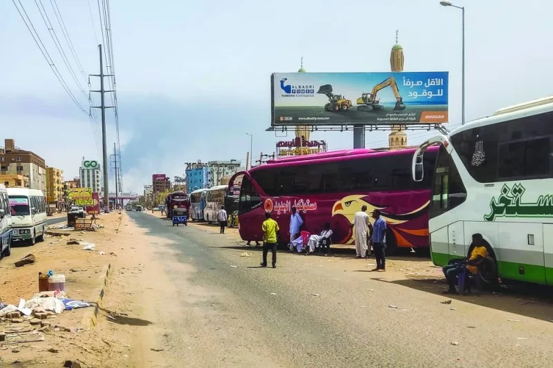 People wait next to passenger buses as smoke billows in an area in Khartoum.