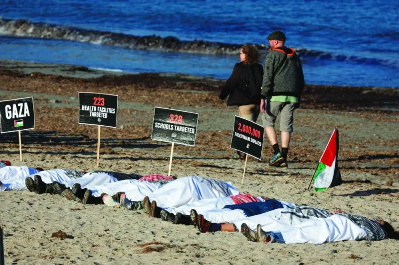 Activists wrapped in white sheets lie in the sand, while placards and a Palestinian flag are displayed during a symbolic protest action at Ghadira Bay in Mellieha, Malta.