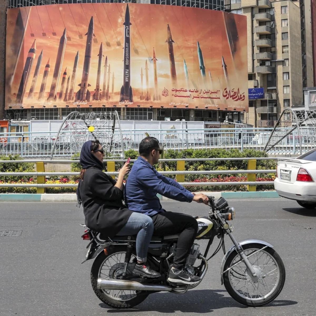 TEHRAN: A woman uses a phone while riding behind a man on a motorcycle moving past a billboard depicting named Iranian ballistic missiles in service, with text in Arabic reading ‘the honest [person’s] promise’ and in Persian ‘Zionist entity is weaker than a spider’s web’, in Valiasr Square in central Tehran on April 15, 2024. – AFP
