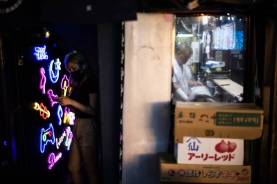 A woman stands on one side of the wall texting in front of a nightclub while, on the other side of the wall, a man works in an izakaya.