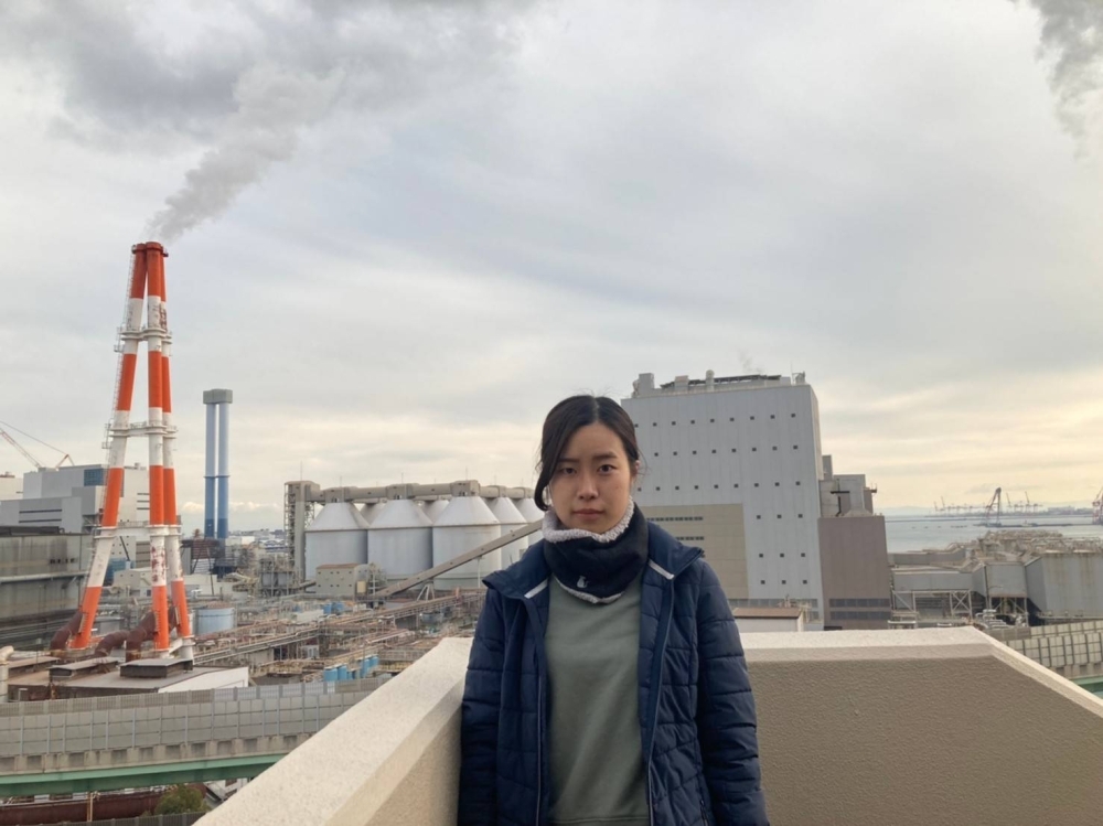 Erina Imai, a plaintiff in a climate lawsuit, poses in front of the coal-fired Kobe Power Plant in Kobe. The red and white tower behind her is private power generation equipment operated by Kobe Steel and is releasing water vapor, not smoke.
