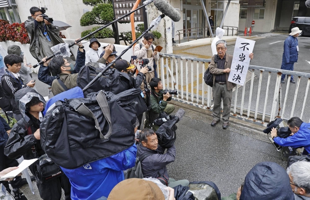 A supporter of Okinawa Gov. Denny Tamaki holds up a sign on Wednesday in front of the Fukuoka District Court's Naha branch saying that the court's ruling — effectively clearing the way for further landfill work to relocate a U.S. base in Okinawa — is "unjust."