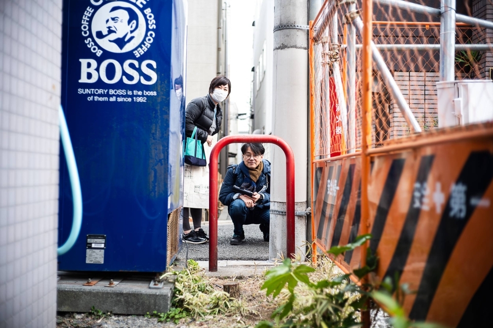 Nama Yoshimura and Hideo Takayama stare through a metal car barrier and down a narrow pathway that likely sits atop an "ankyo," or subterranean waterway, in Tokyo’s Koenji neighborhood.