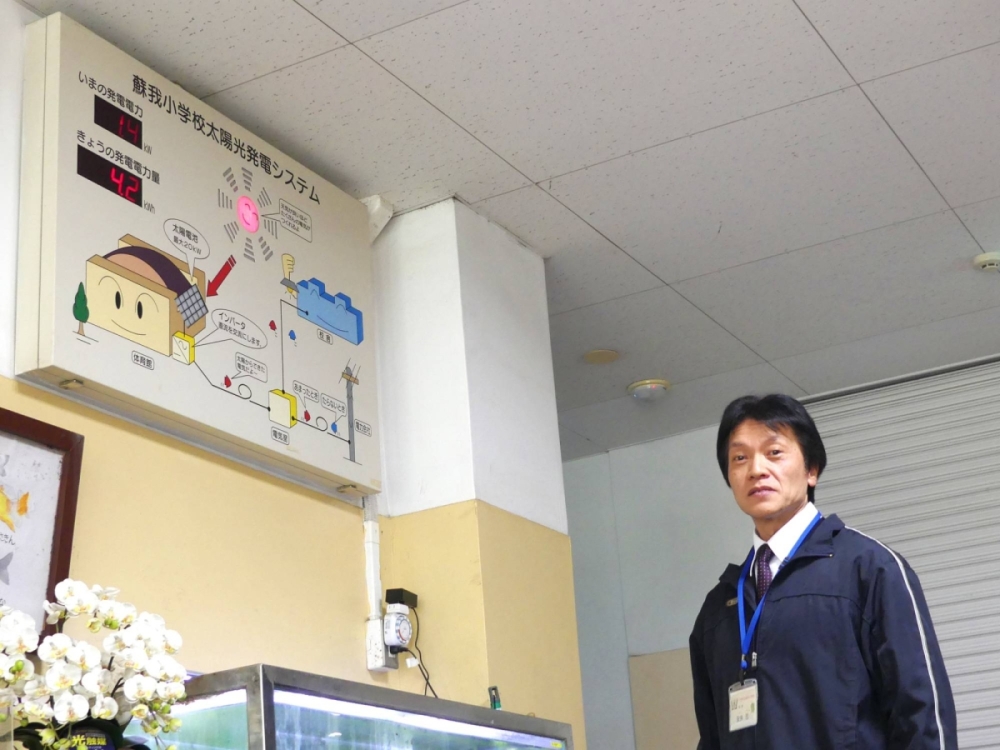 Soga Elementary School Principal Shinobu Yasui stands next to a monitor partially showing the amount of solar power generated on a real-time basis. A more advanced monitoring system is used now that all solar panels have been installed. Soga Elementary School Principal Shinobu Yasui stands next to a monitor partially showing the amount of solar power generated on a real-time basis. A more advanced monitoring system is used now that all solar panels have been installed.