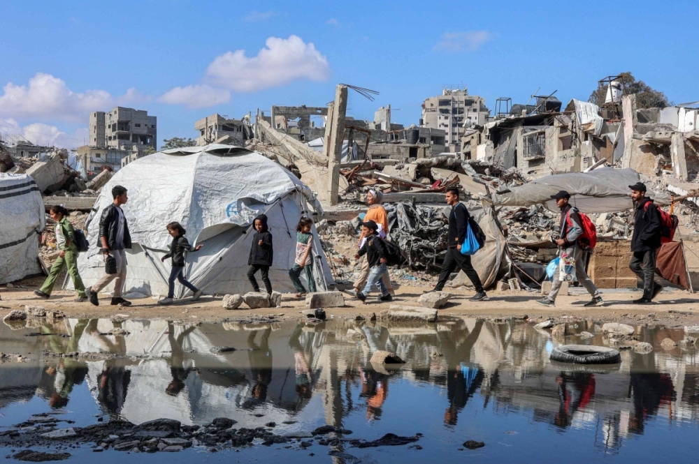 People walk past a puddle of water by a tent shelter erected near the rubble of a collapsed building in the Nasr neighborhood in western Gaza on Tuesday. People walk past a puddle of water by a tent shelter erected near the rubble of a collapsed building in the Nasr neighborhood in western Gaza on Tuesday.
